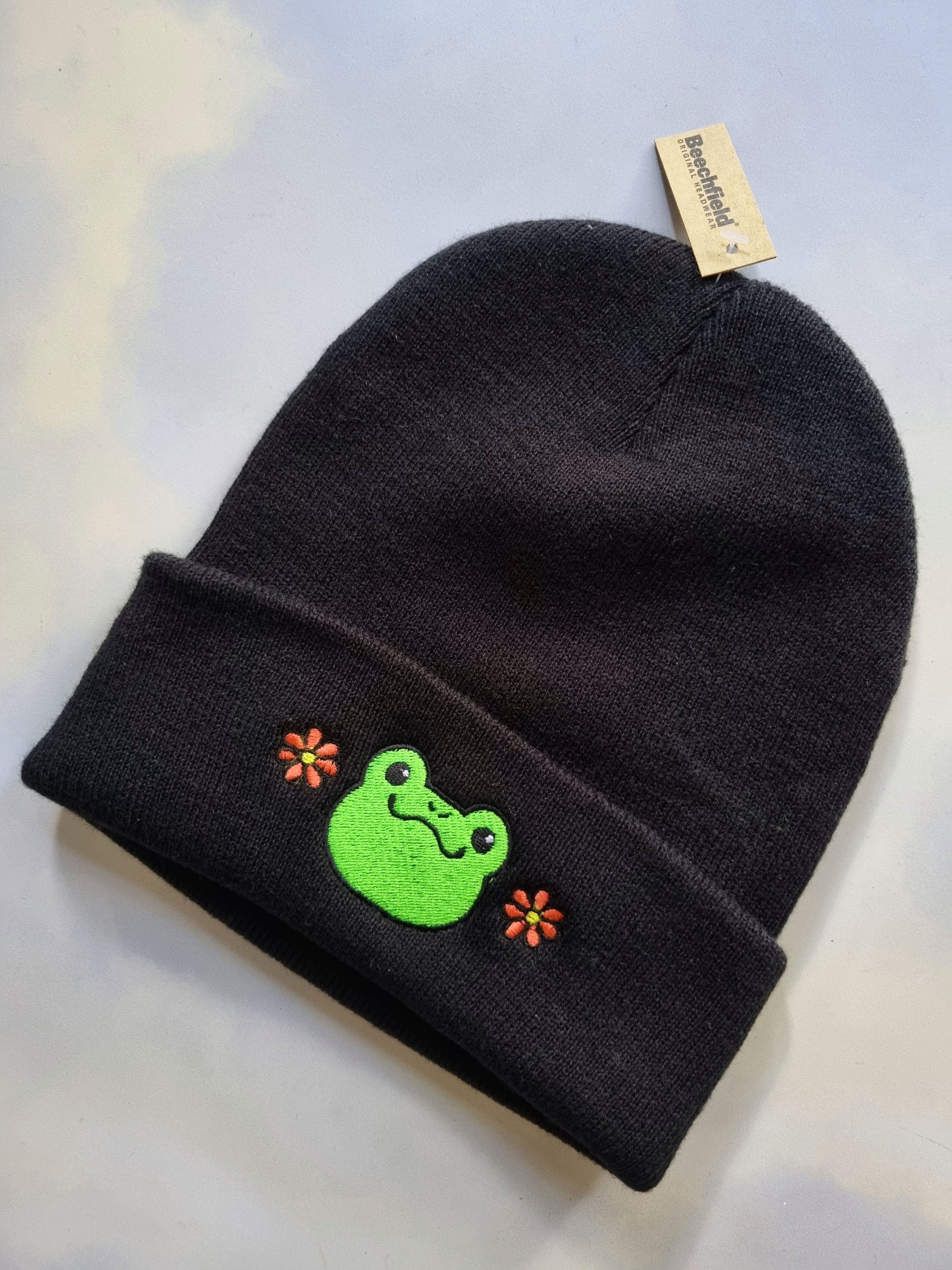 Black beanie with a green frog design and flower accents on a light background