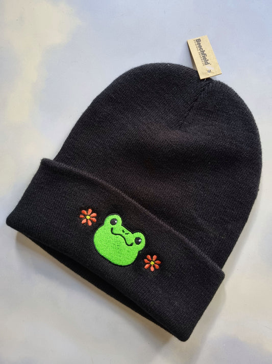 Black beanie with a green frog design and flower accents on a light background