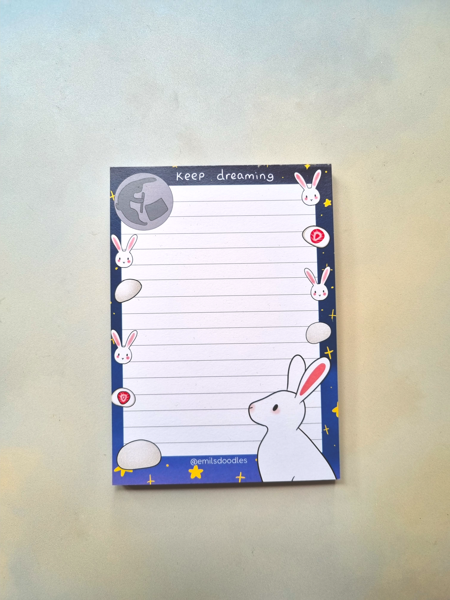 A lined notepad with the words "keep dreaming" at the top. It is decorated with rabbits. Lower right corner is a rabbit and opposite in the top left is a moon with a rabbit pounding mochi.